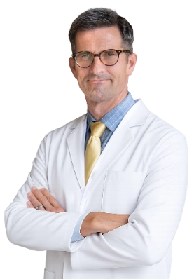 Dr. Joshua Kindelan is captured in a professional portrait wearing a white lab coat over a blue plaid dress shirt and a yellow tie. He is standing with his arms crossed in a clinical environment.